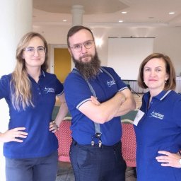 Circinus Training Center Adam Pluciński - Trzy osoby w niebieskich koszulkach polo z logo firmy szkoleniowej, stojące w sali konferencyjnej z projektorem i stołami w tle. Mężczyzna z brodą i w szelkach.