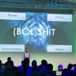 Prelegent w okularach i muchą na scenie prezentuje slajd z napisem '(BÓL)SHIT' otoczonym słowami Rynek, Firma, Telemarketer, Klient, podczas szkolenia dla publiczności.