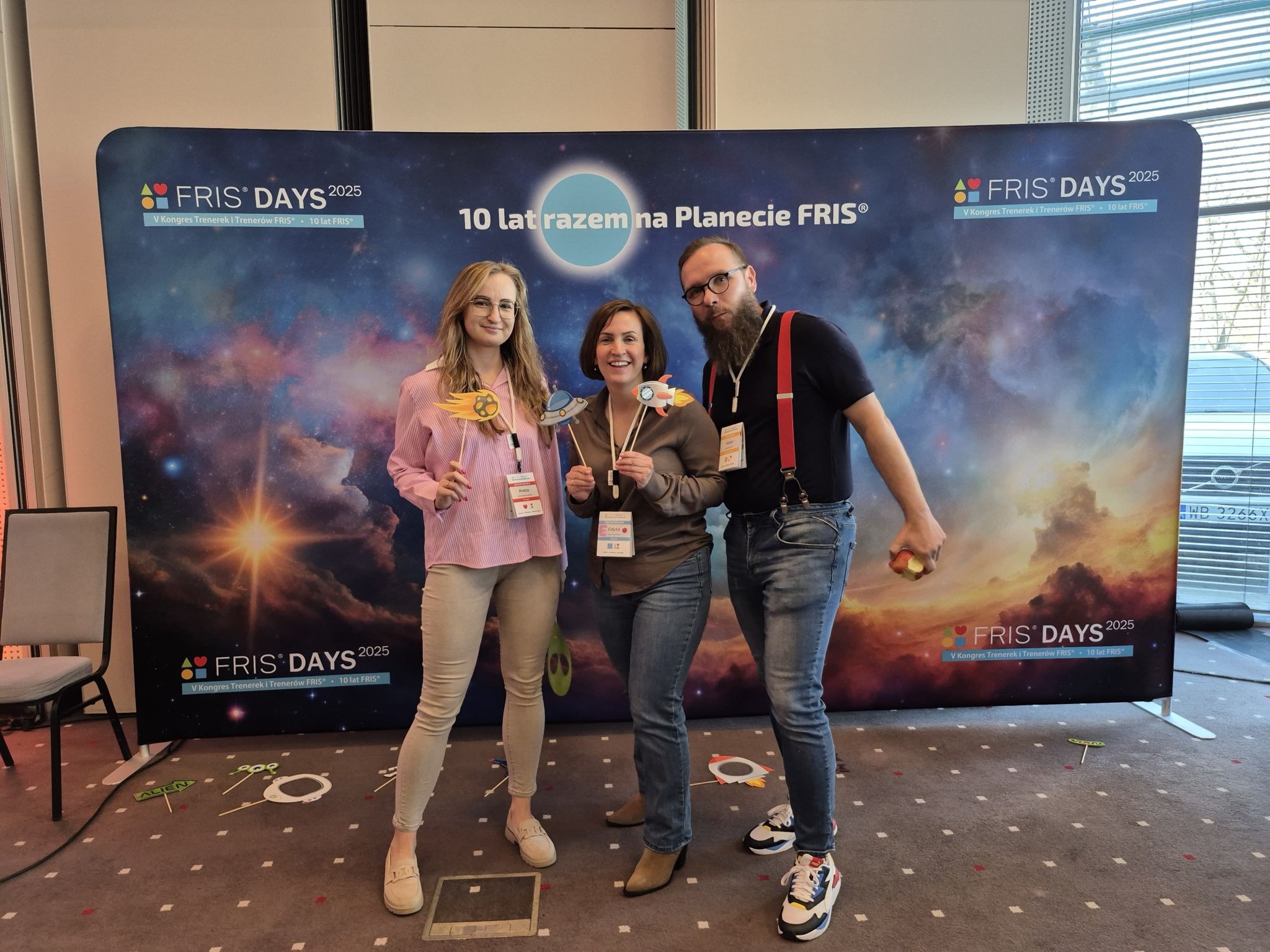 Trzy osoby pozują z rekwizytami na tle baneru 'FRIS Days 2025'. Uczestnicy konferencji z kosmicznymi gadżetami. Uśmiechnięte twarze, wesoła atmosfera.