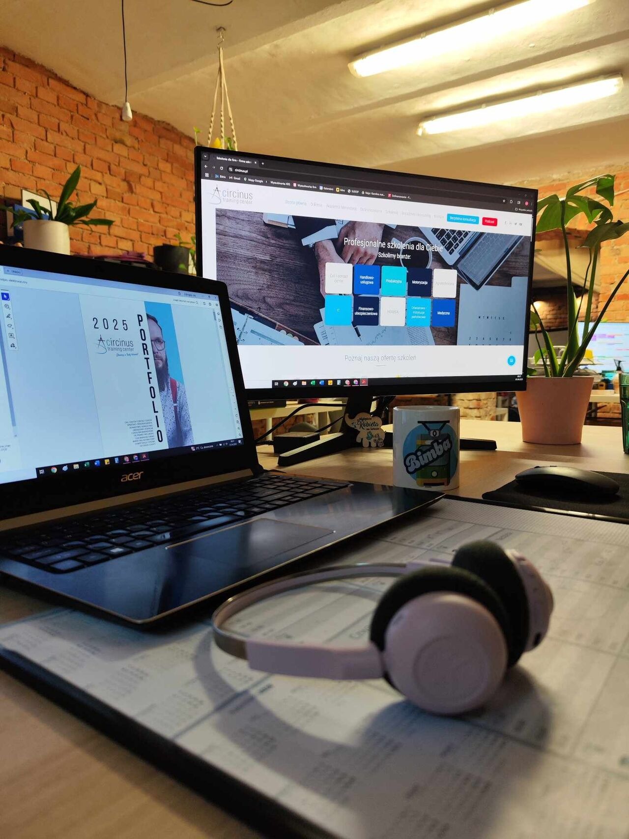 Biurko z dwoma monitorami, laptopem z otwartym portfolio firmy szkoleniowej Circinus, słuchawkami i kubkiem. Widoczny fragment ceglanej ściany i rośliny doniczkowe.