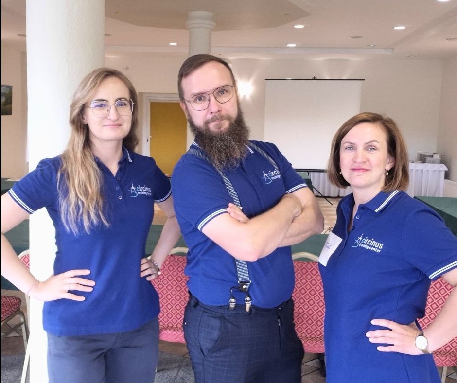 Trzy osoby w niebieskich koszulkach polo z logo firmy szkoleniowej, stojące w sali konferencyjnej z projektorem i stołami w tle. Mężczyzna z brodą i w szelkach.