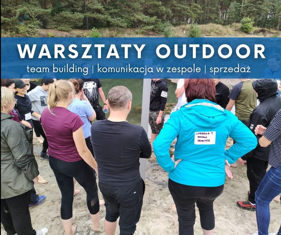 Warsztaty outdoor: grupa osób na piasku, w tle las. Uczestnicy w trakcie ćwiczenia team building, komunikacja w zespole i sprzedaż. Widoczny napis na bluzie.