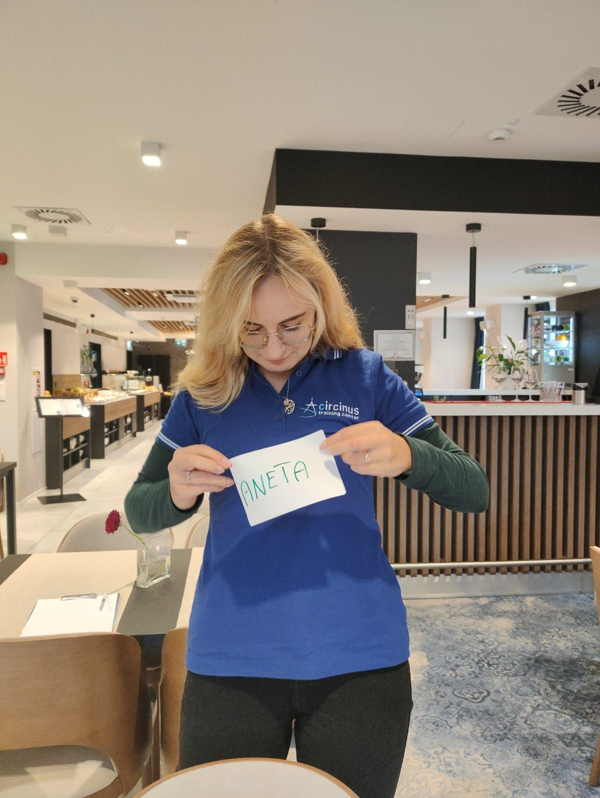 Kobieta w niebieskiej koszulce z logo 'Circinus training center' trzyma kartkę z napisem 'ANETA' w jasnym wnętrzu restauracji.