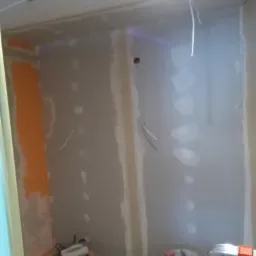 Wnętrze w trakcie prac przygotowawczych: ściany z płyt kartonowo-gipsowych, szpachlowane łączenia, widoczne przewody elektryczne, fragment pomalowanej na pomarańczowo ściany, narzędzia budowlane...