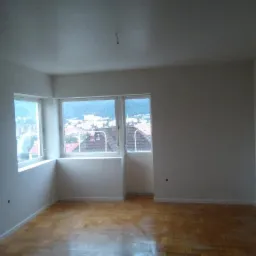 Pusty pokój po odświeżeniu ścian, widoczne drewniane parkiety, białe ściany i okna z widokiem na miasto.