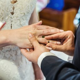 Justart-Studio - Moment zakładania złotej obrączki na palec panny młodej w koronkowej sukni ślubnej, zbliżenie dłoni.