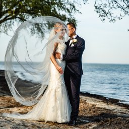 Justart-Studio - Para młoda na plaży, panna młoda w koronkowej sukni z welonem powiewającym na wietrze, pan młody w granatowym garniturze, w tle morze i drzewa.