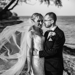 Justart-Studio - Czarno-białe zdjęcie pary młodej na tle jeziora, panna młoda w koronkowej sukni, welon powiewa na wietrze, pan młody w garniturze z muchą.