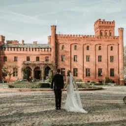 Para młoda idzie w kierunku ceglanego zamku w słoneczny dzień, suknia panny młodej z długim trenem.