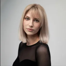 Portret kobiety z blond włosami, delikatnym makijażem i czarną, prześwitującą bluzką na jasnym tle.