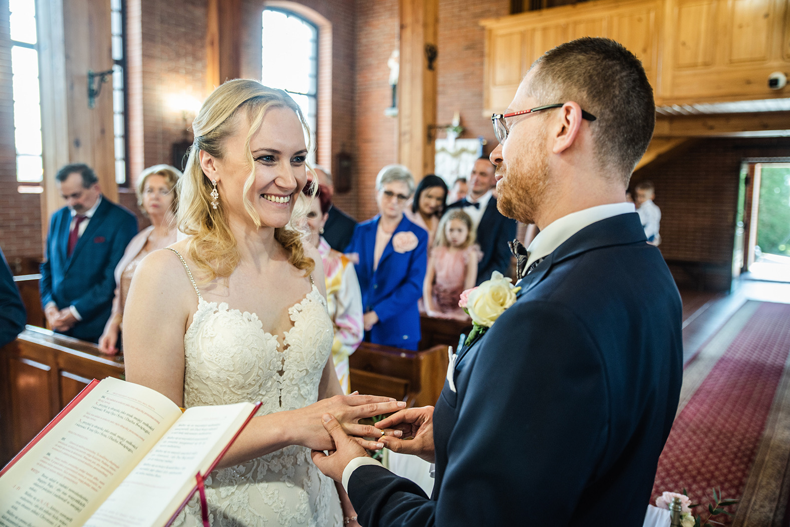 Panna młoda z blond włosami uśmiecha się podczas ceremonii ślubnej, gdy pan młody zakłada jej obrączkę w kościele z ceglanymi ścianami, w tle goście.