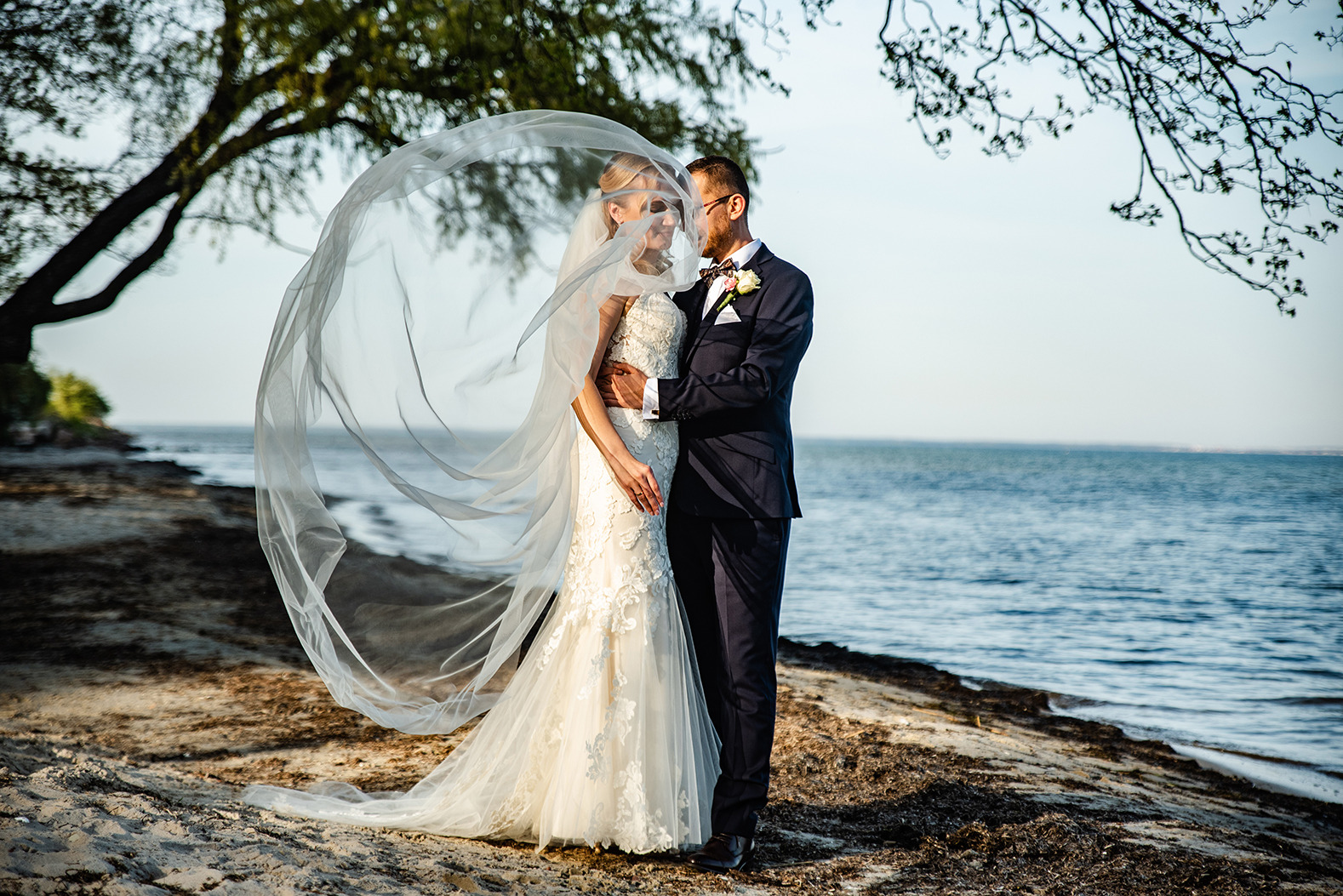 Para młoda na plaży, panna młoda w koronkowej sukni z welonem powiewającym na wietrze, pan młody w granatowym garniturze, w tle morze i drzewa.