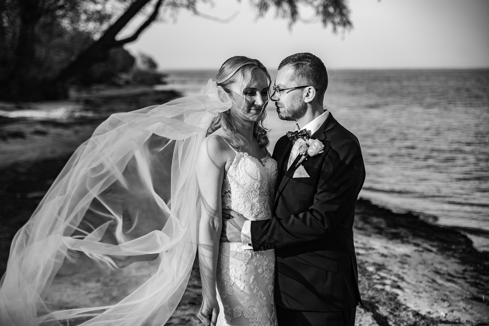 Czarno-białe zdjęcie pary młodej na tle jeziora, panna młoda w koronkowej sukni, welon powiewa na wietrze, pan młody w garniturze z muchą.