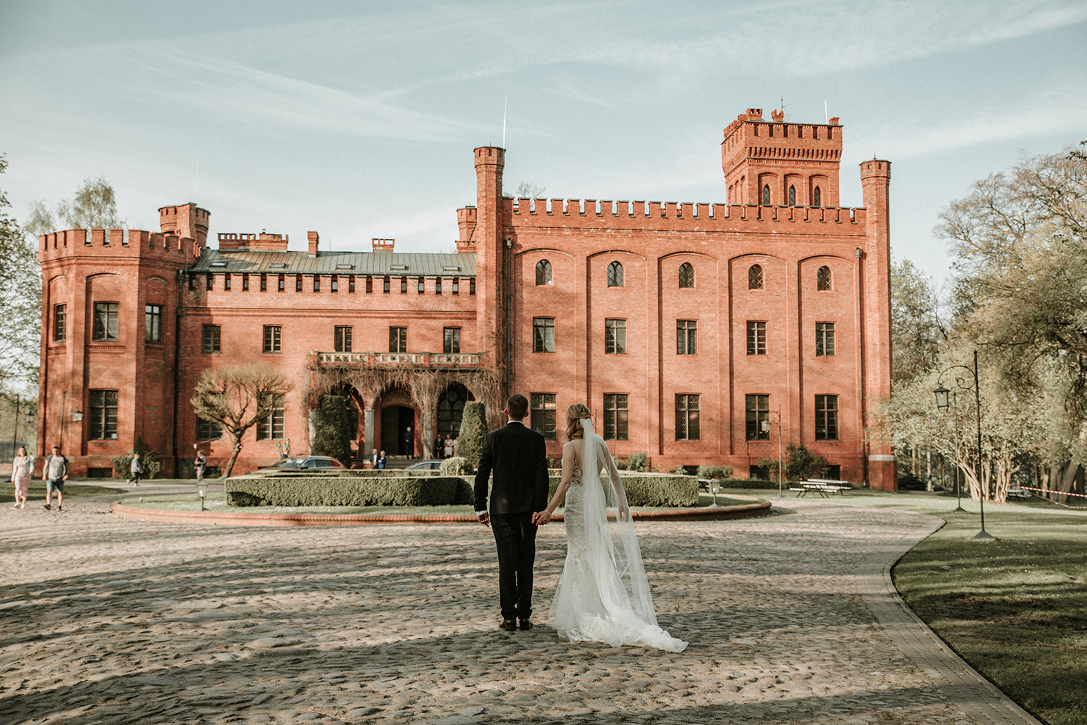 Para młoda idzie w kierunku ceglanego zamku w słoneczny dzień, suknia panny młodej z długim trenem.