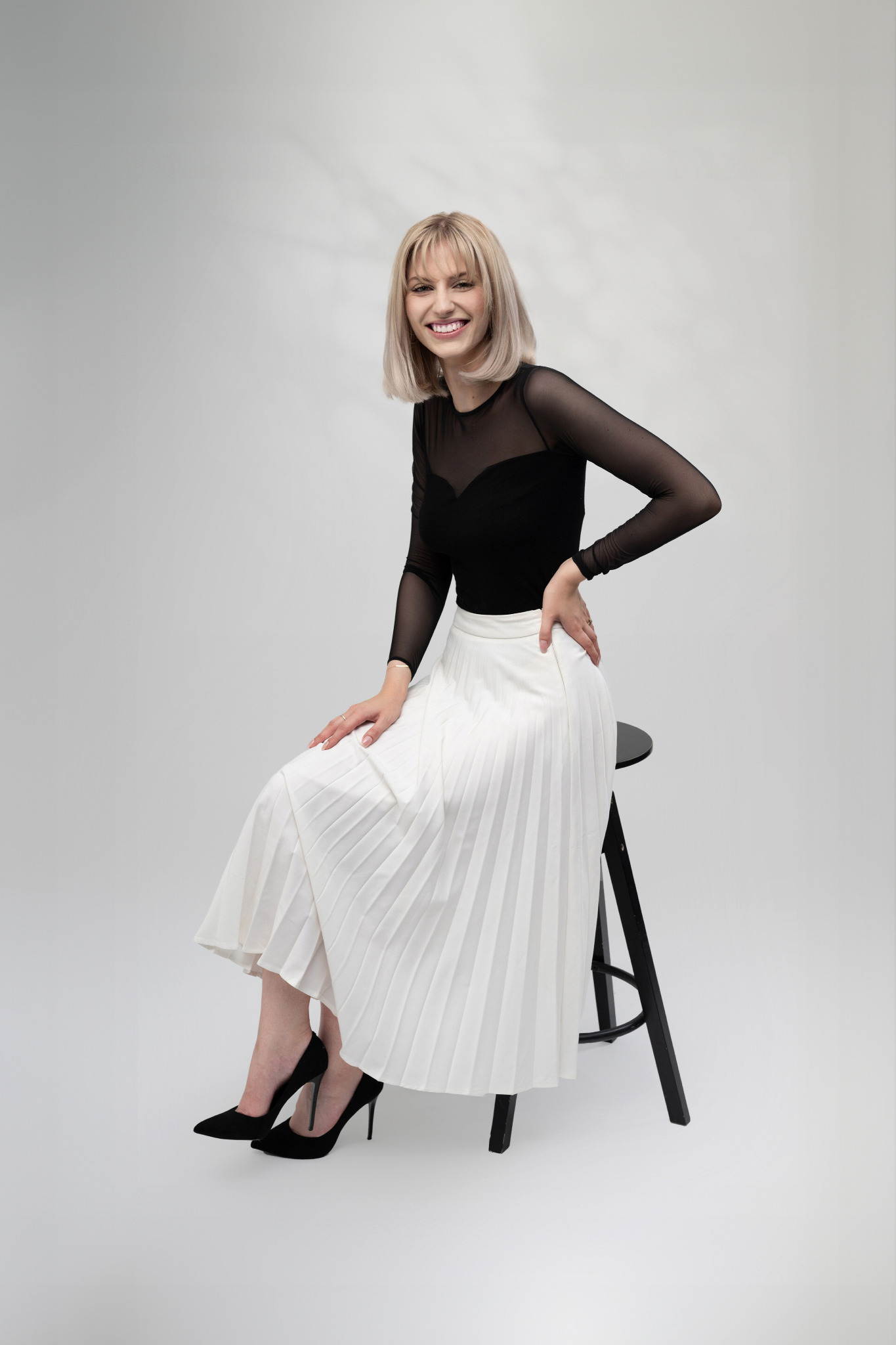 Uśmiechnięta blondynka w czarnej bluzce z siateczki i białej plisowanej spódnicy, siedząca na czarnym stołku barowym na jasnym tle, pozująca w czarnych szpilkach.