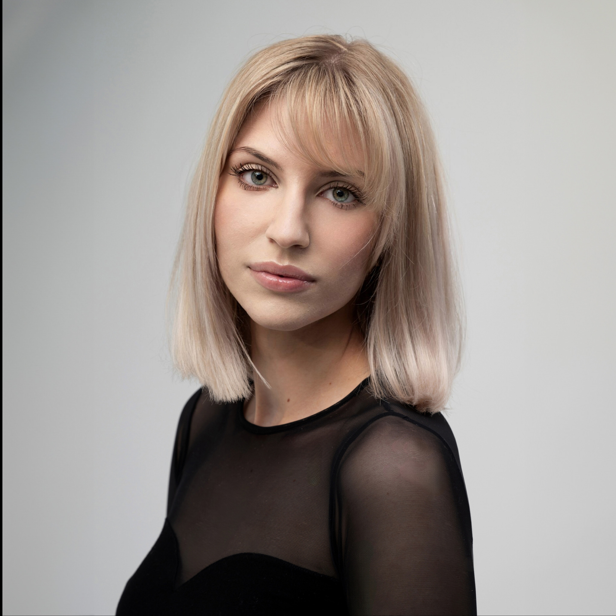 Portret kobiety z blond włosami, delikatnym makijażem i czarną, prześwitującą bluzką na jasnym tle.