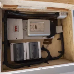 Otwarta skrzynka z licznikiem elektrycznym, bezpiecznikami i przewodami w czarnej karbowanej osłonie, widok z góry.