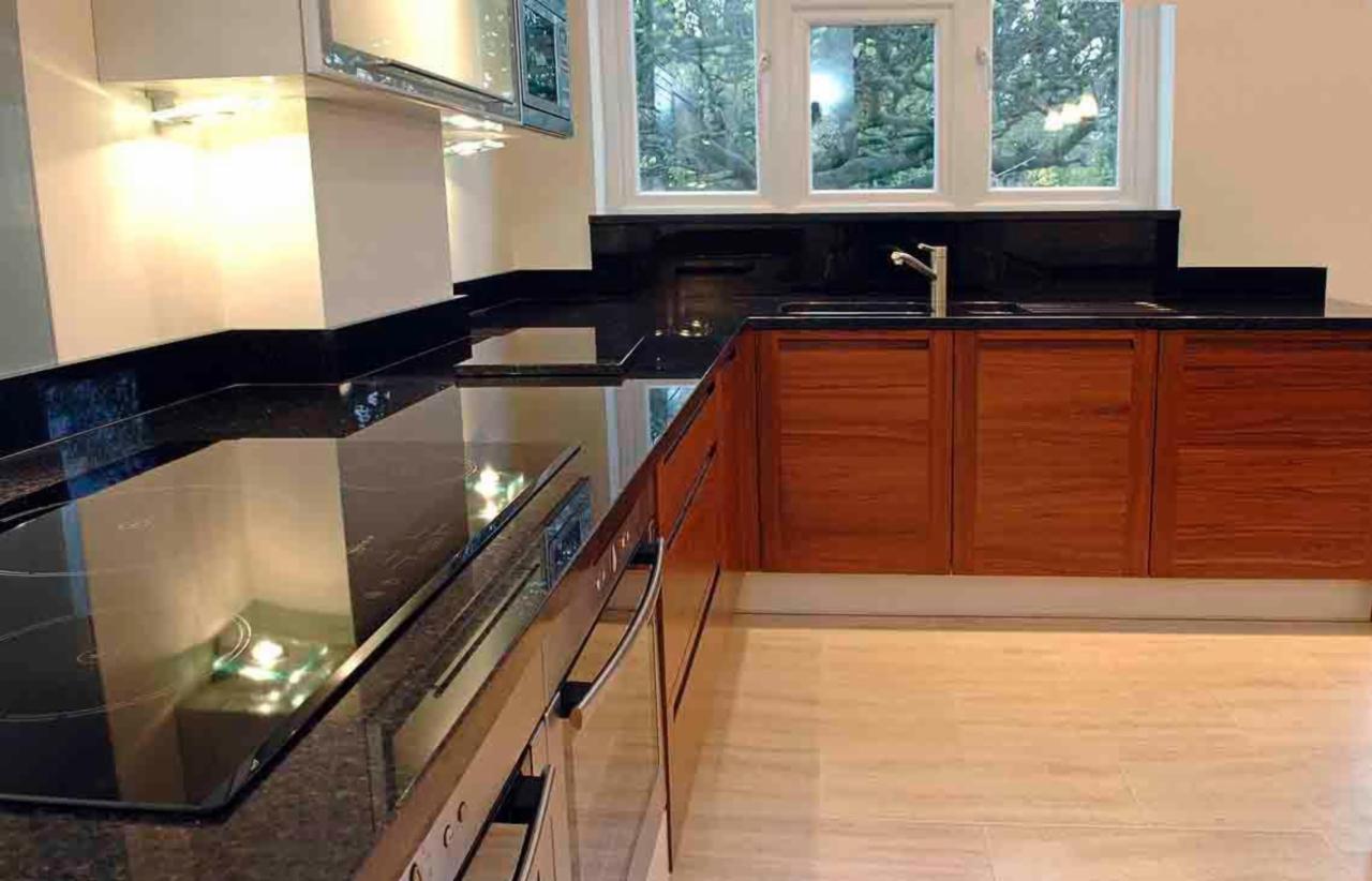 Ciemny, błyszczący blat kuchenny z kamienia, z wbudowaną płytą indukcyjną, widok na drewniane szafki i okno z widokiem na drzewa.