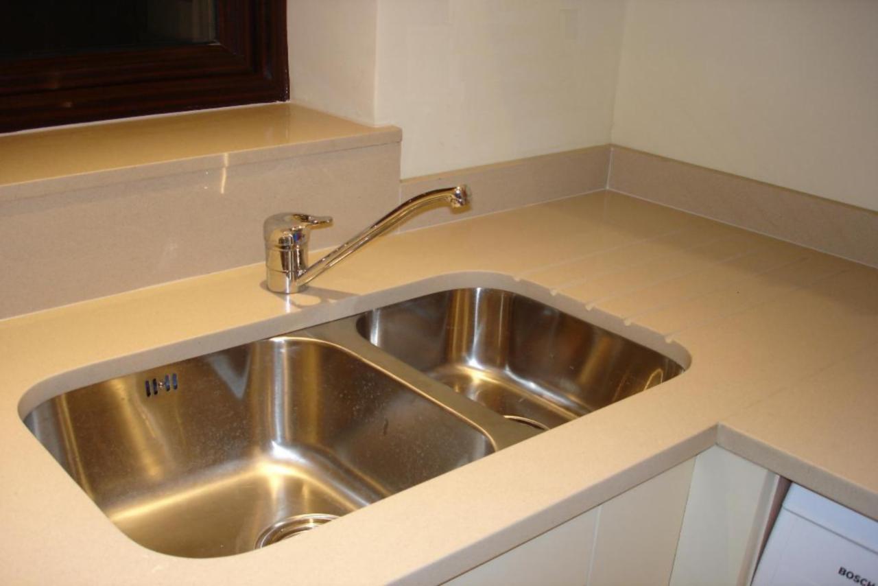 Beżowy blat kamienny z podwójnym zlewem ze stali nierdzewnej i chromowaną baterią, widoczne delikatne wzory w kamieniu, oświetlenie górne.