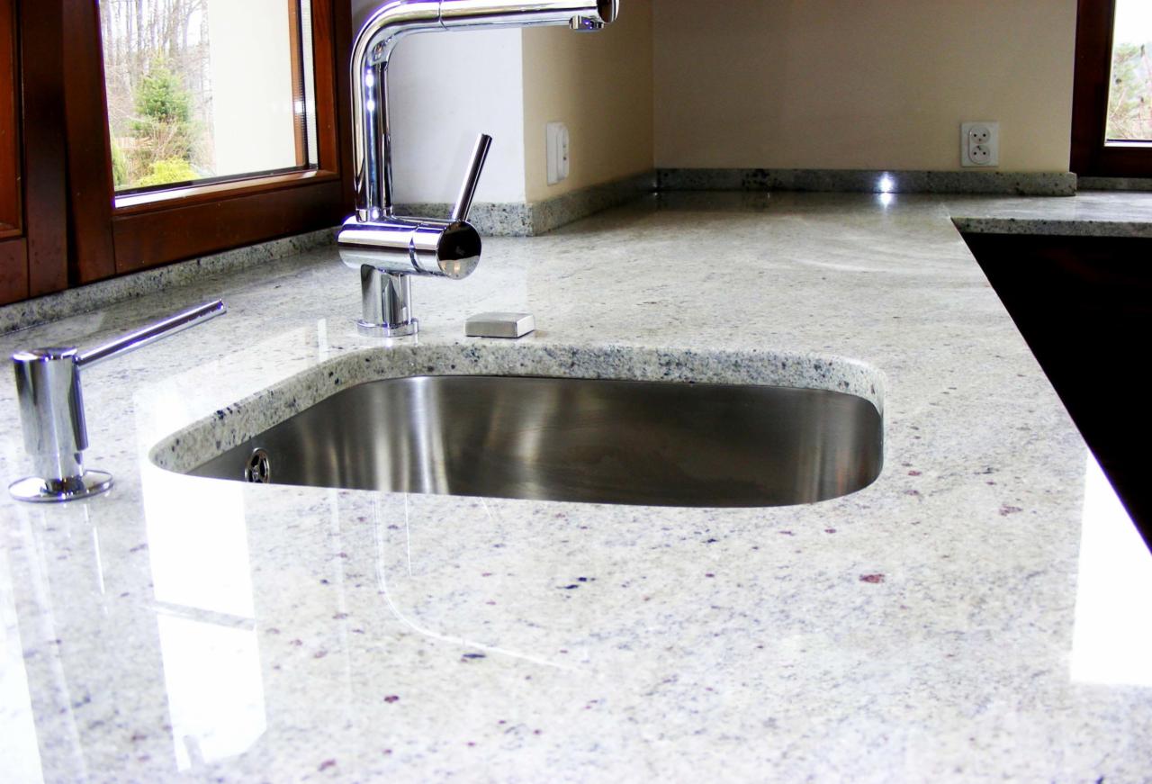 Biały blat kuchenny z delikatnym wzorem, z wbudowanym zlewem ze stali nierdzewnej i chromowaną baterią, uzupełniony dozownikiem mydła i małą gąbką, w tle okno z drewnianą ramą i fragment ściany...