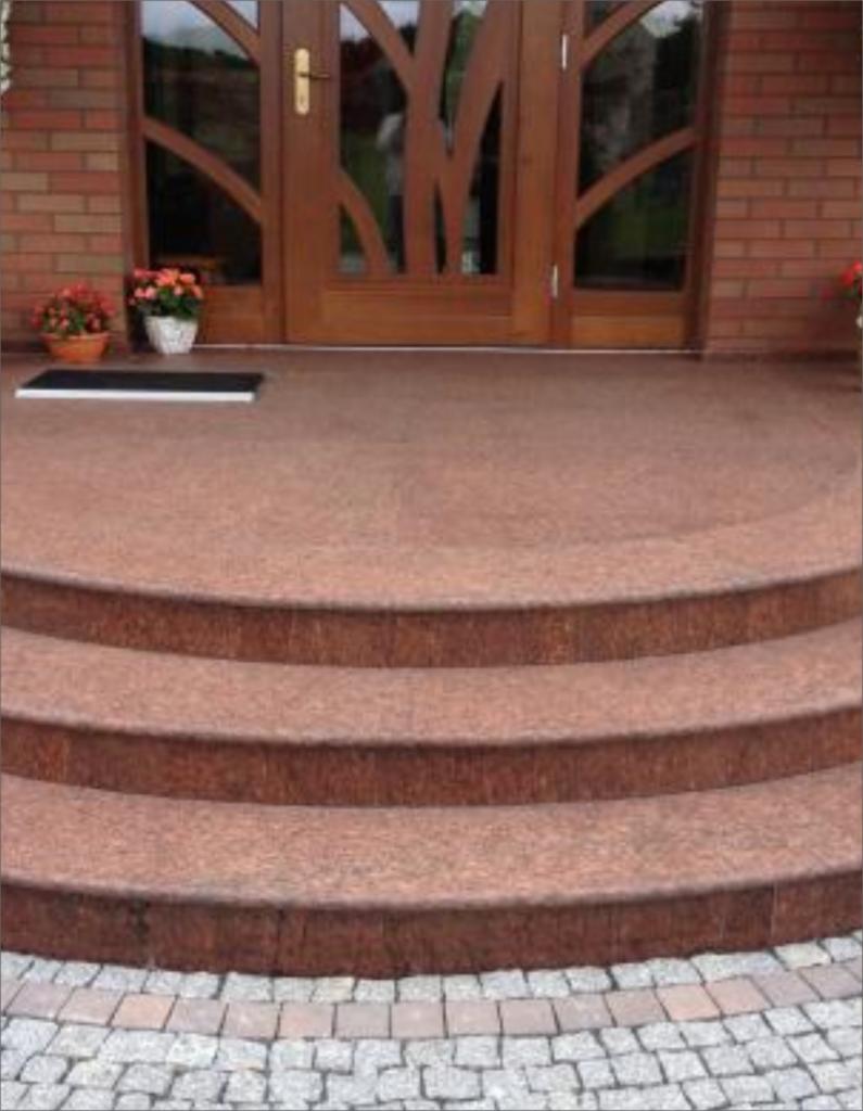 Zewnętrzne, półokrągłe schody wejściowe z czerwono-brązowego kamienia, wykończone kostką brukową, prowadzące do drewnianych drzwi z dekoracyjnym szkleniem.