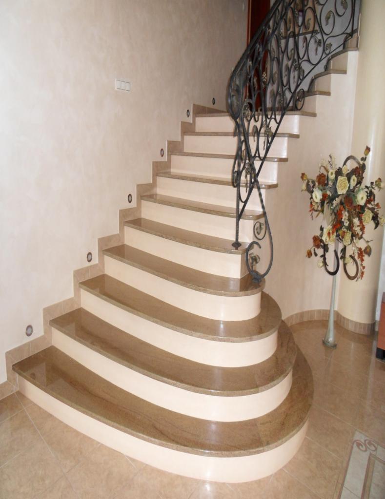 Kamienne schody z beżowymi stopniami i ozdobną, kutą balustradą, oświetlone punktowymi lampami w ścianie, z widocznym wazonem z kwiatami.