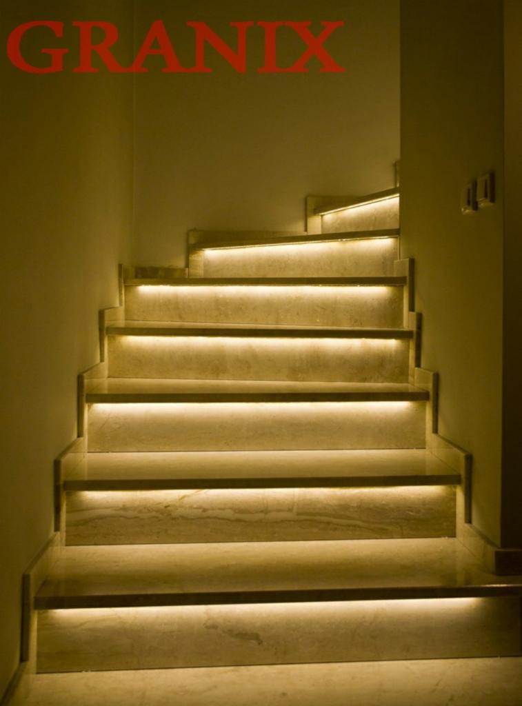 Bieg schodów z podświetleniem LED pod każdym stopniem, wykonanych z jasnego kamienia z delikatnym wzorem żyłkowania, widoczne z dołu do góry, logo firmy w górnym lewym rogu.