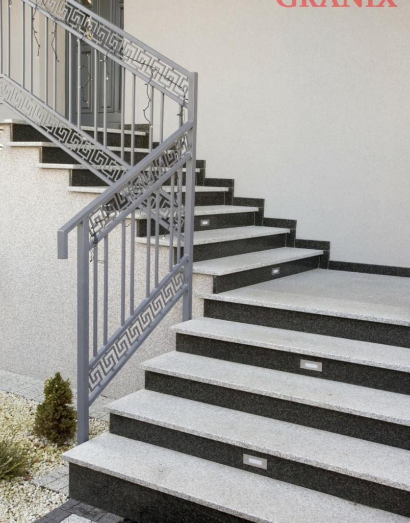 Zewnętrzne schody z szarego i czarnego kamienia z metalową balustradą zdobioną greckim wzorem, oświetlone małymi lampkami w stopniach.