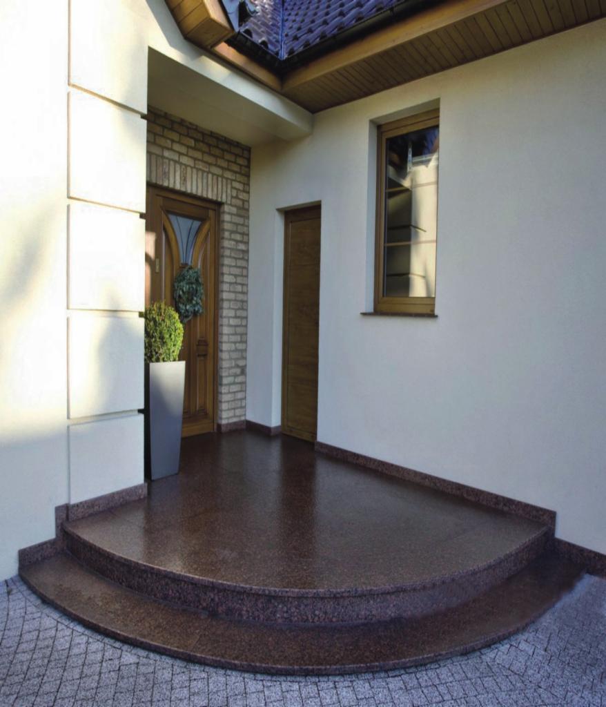 Wejście do domu z półokrągłymi schodami i podestem wyłożonymi brązowym kamieniem z widoczną strukturą, na zewnątrz kostka brukowa, widoczne drzwi wejściowe i okno.
