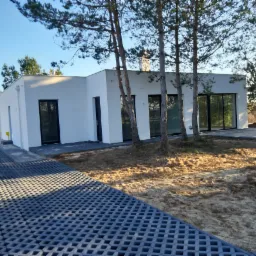 Nowoczesny dom jednorodzinny z białymi ścianami i czarnymi oknami, otoczony młodymi drzewami iglastymi, z podjazdem wyłożonym ażurowymi płytami betonowymi.