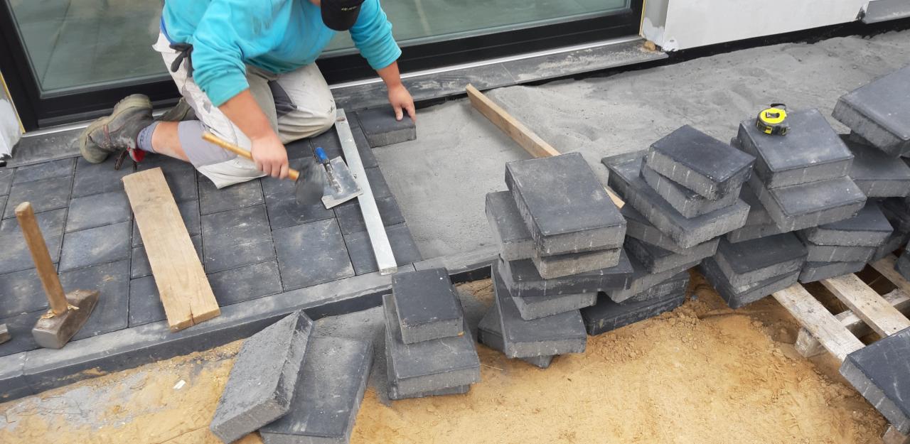 Pracownik w trakcie układania szarej kostki brukowej na podsypce piaskowej, widoczne narzędzia: młotek, poziomica, łata, miarka.