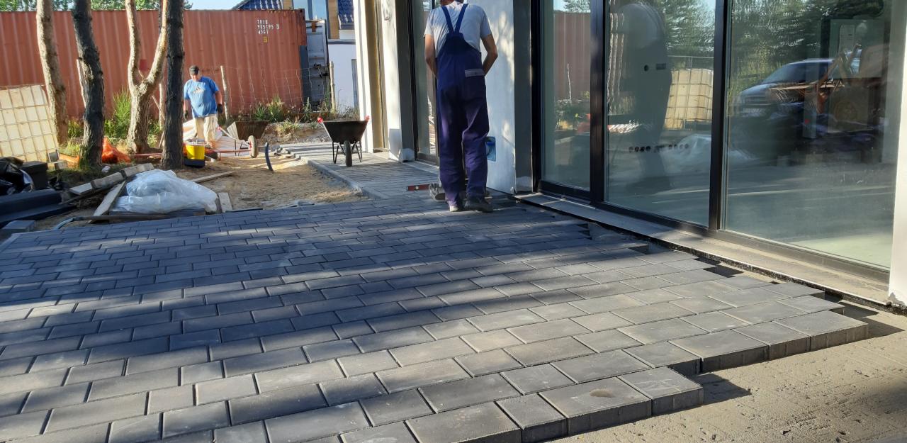 Montaż szarej kostki brukowej prostokątnej na podjeździe, widoczny pracownik w kombinezonie oraz taczka i narzędzia w tle.