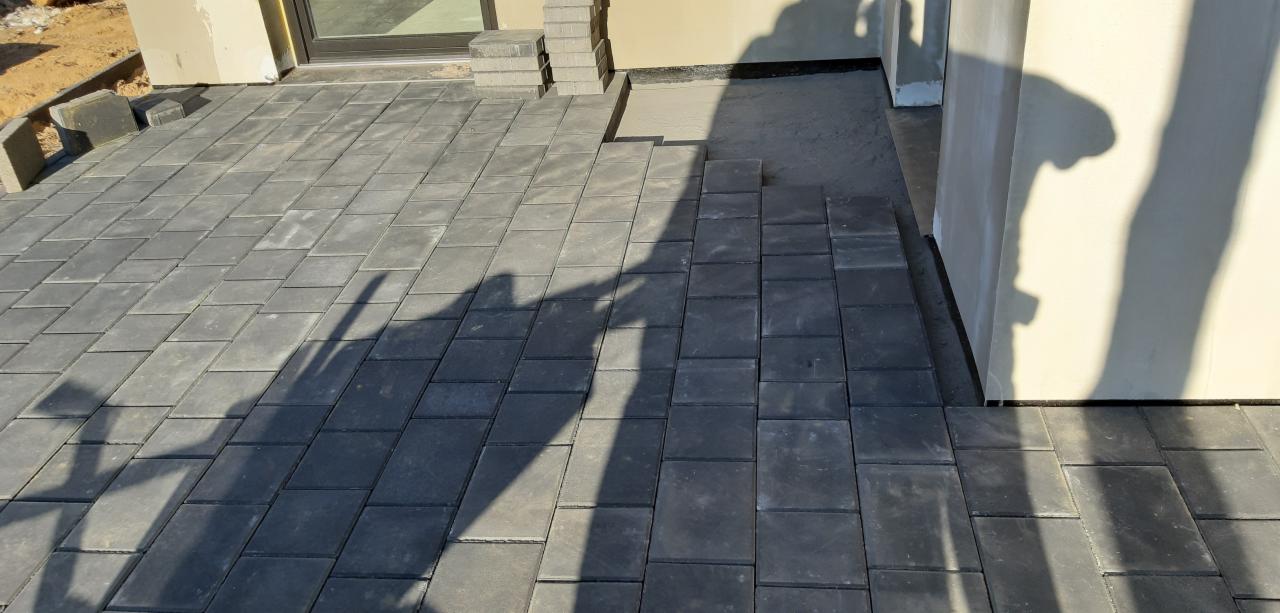 Częściowo ułożona, grafitowa kostka brukowa na podjeździe, widoczne zapasy kostki obok budynku, cień osoby fotografującej.