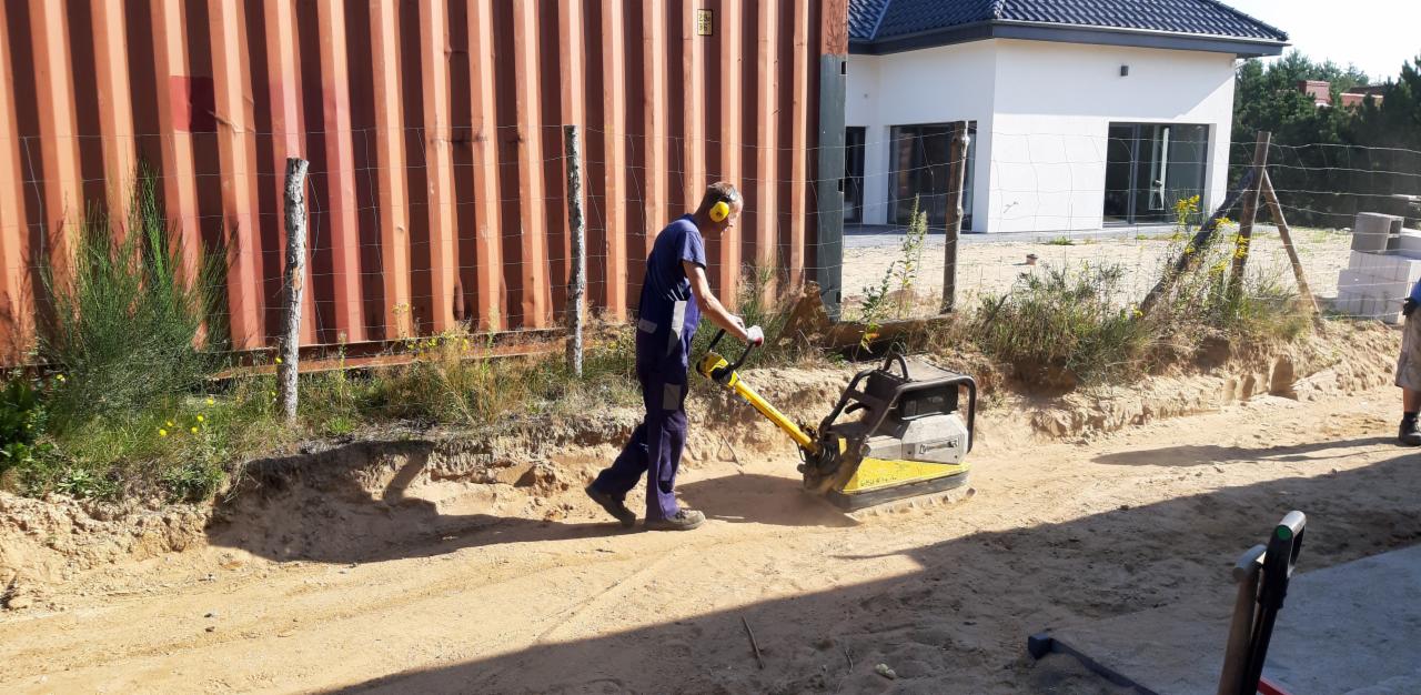 Pracownik w niebieskim kombinezonie ubija piasek zagęszczarką wibracyjną, przygotowując podłoże pod przyszłą nawierzchnię, z widocznym kontenerem i domem w tle.