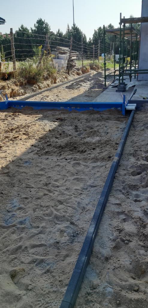 Przygotowanie podłoża pod kostkę brukową: wyrównany teren z piaskiem, ustawione szyny prowadzące i niebieska listwa do wyrównywania.