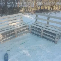 Dwa surowe, drewniane siedziska ogrodowe wykonane z palet, ustawione na śniegu w słoneczny dzień. Widoczna butelka oleju obok jednego z siedzisk.
