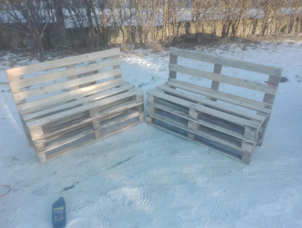 Dwa surowe, drewniane siedziska ogrodowe wykonane z palet, ustawione na śniegu w słoneczny dzień. Widoczna butelka oleju obok jednego z siedzisk.