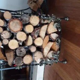 Kute metalowe stanowisko na drewno kominkowe, wypełnione polanami o różnej średnicy, na tle drewnianej podłogi i fragmentu marmurowego kominka.