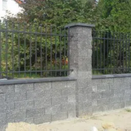Nowoczesne ogrodzenie z szarej kostki betonowej i metalowych prętów, z widocznym fragmentem ulicy i stertą piasku obok.