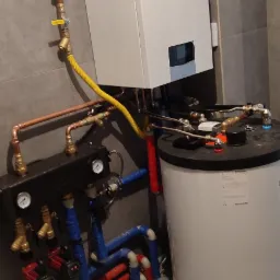 Kocioł grzewczy Vaillant z podłączeniami hydraulicznymi i zasobnikiem ciepłej wody w szarej łazience z płytkami.
