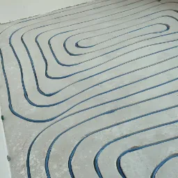 Niebieskie rurki układane w spiralne wzory w szarej wylewce podłogowej, przygotowane do zalania betonem.