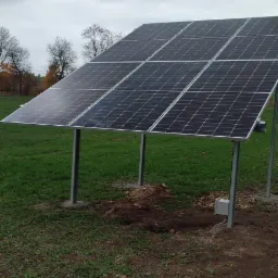 Panel fotowoltaiczny na metalowych słupkach, zamontowany na trawiastym terenie z widoczną ziemią wokół słupków, pochmurne niebo w tle.