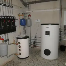 Zestaw urządzeń grzewczych w kotłowni: dwa białe zasobniki, rozdzielacz z manometrami, naczynia wzbiorcze oraz wiszący piec gazowy, rury miedziane łączące elementy.