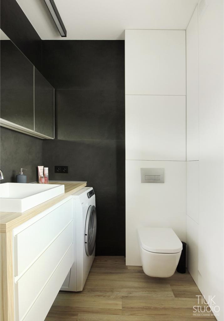 Nowoczesna łazienka z czarną ścianą, białą zabudową meblową z umywalką nablatową, pralką i podwieszaną toaletą na drewnopodobnej podłodze.