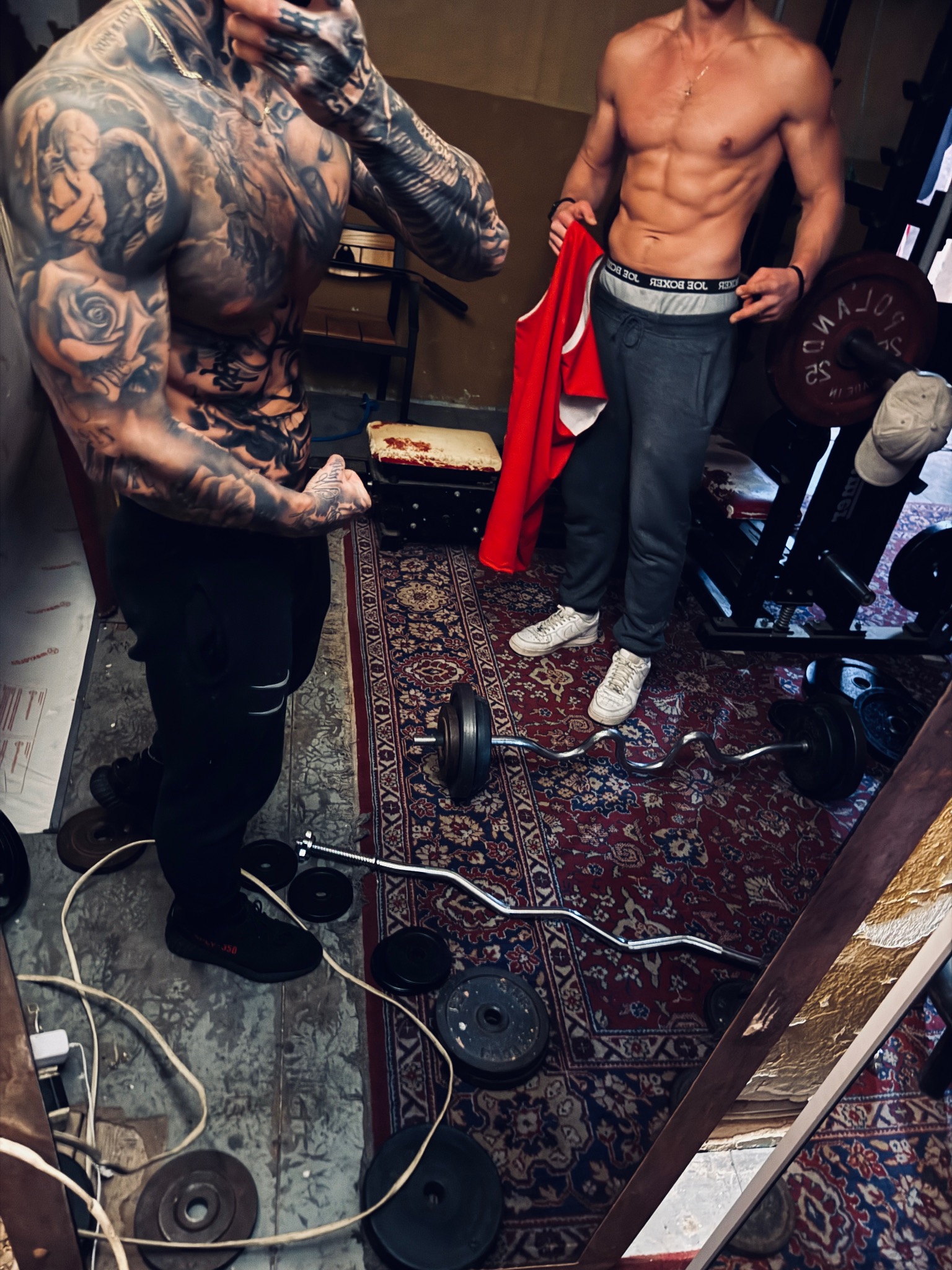 Dwóch wysportowanych mężczyzn w domowej siłowni. Jeden z tatuażami na całym ciele, drugi z widocznymi mięśniami brzucha. W tle sprzęt do ćwiczeń i dywan.