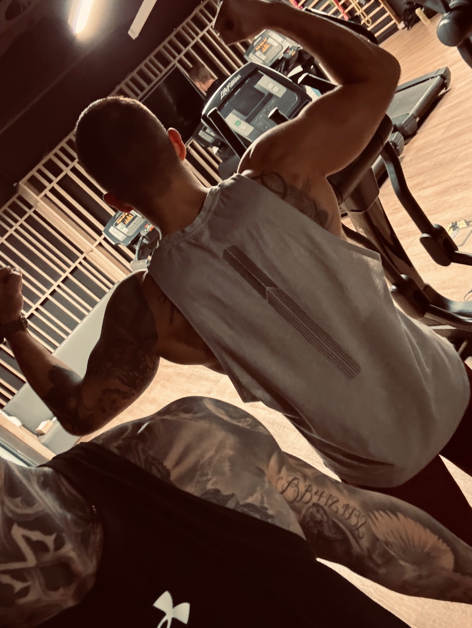 Mężczyzna z tatuażami, w szarej koszulce, prezentuje muskulaturę bicepsów na tle sprzętu do ćwiczeń w siłowni. Widoczny fragment bieżni i maszyny eliptycznej.