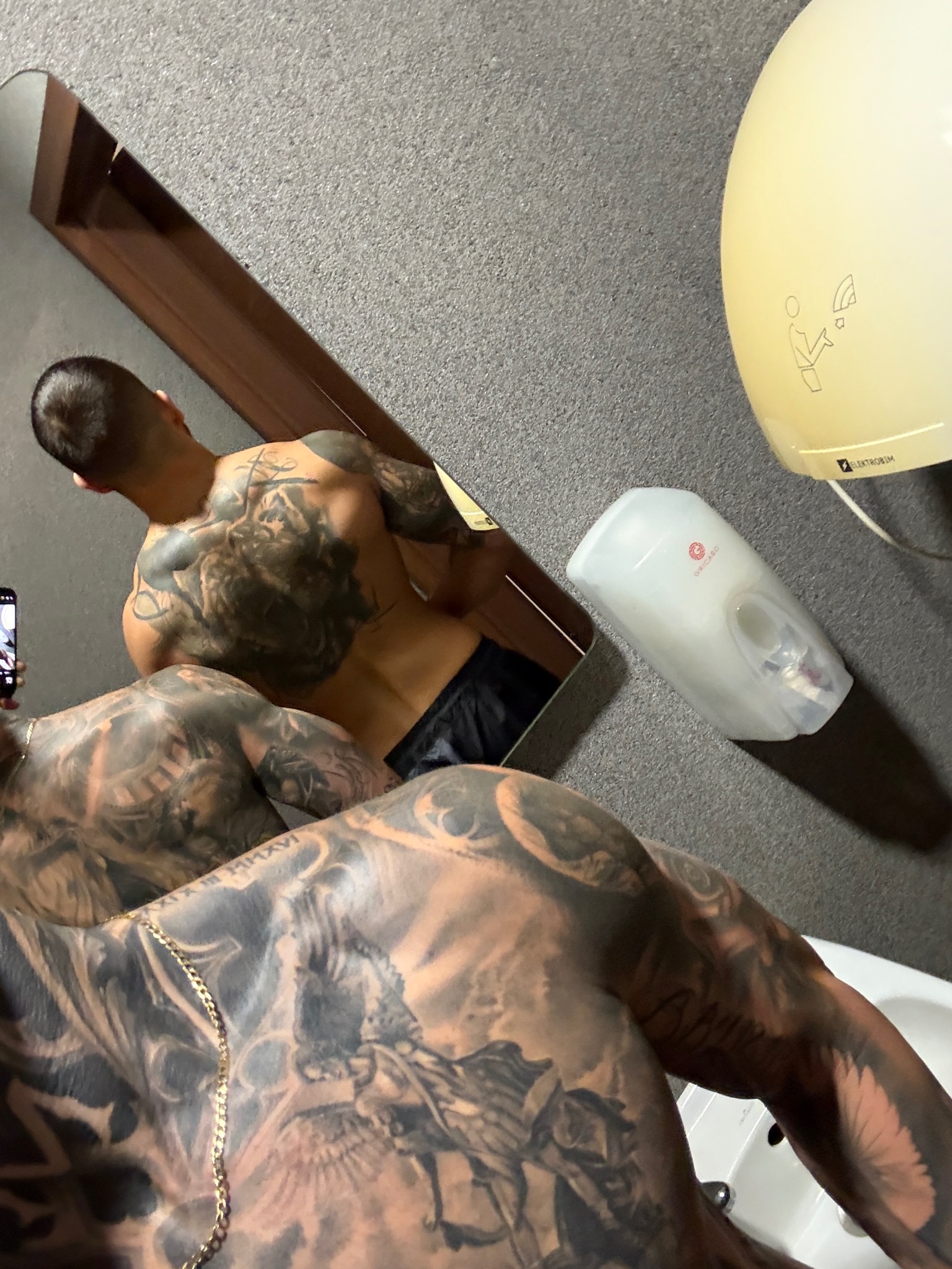 Mężczyzna z tatuażami prezentuje muskulaturę, pozując do zdjęcia w lustrze. Widoczny fragment łazienki z dozownikiem mydła i suszarką do rąk.