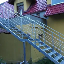 Zewnętrzne schody metalowe z balustradą z rurek, prowadzące do drzwi na piętrze budynku o żółtej elewacji i czerwonym dachu.