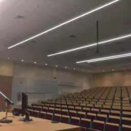 Sala audytoryjna z rzędami krzeseł, oświetlona liniowymi lampami LED, widoczny pulpit prelegenta z mikrofonem i monitorem.