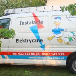 Biały samochód dostawczy z napisem 'Instalacje Elektryczne', numerem telefonu i adresem strony internetowej, ozdobiony rysunkami Kaczora Donalda i jego siostrzeńców, zaparkowany na trawie...